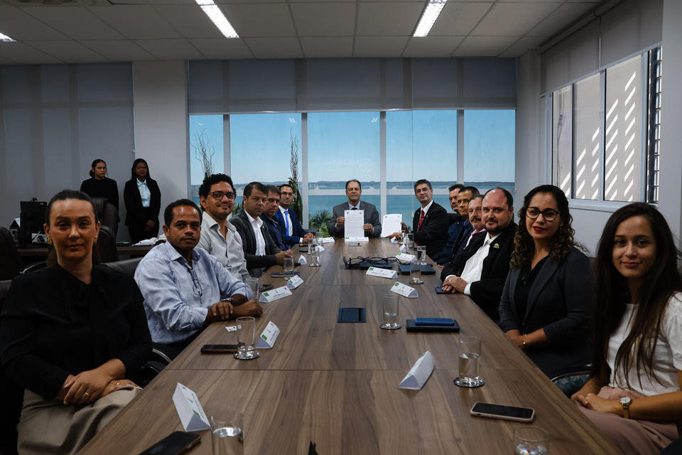 Mesa comprida com vários homens e mulheres sentados e janela de vidro ao fundo